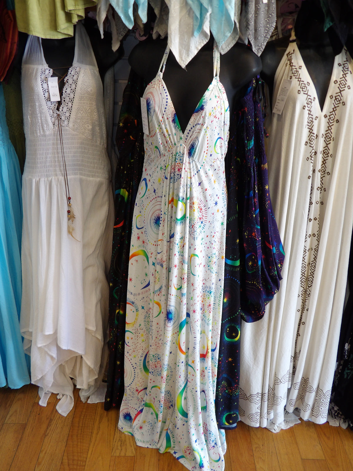 A sleeveless white halter maxi dress with a colorful rainbow and celestial print is displayed on a black mannequin in a clothing store. Other dresses are visible on mannequins in the background.