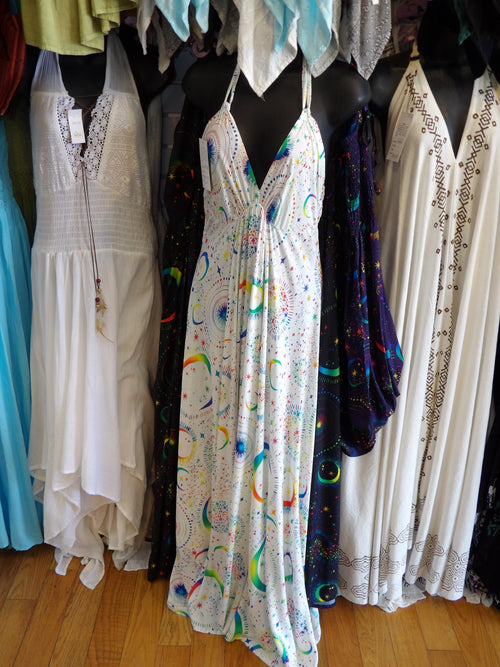 A sleeveless white halter maxi dress with a colorful rainbow and celestial print is displayed on a black mannequin in a clothing store. Other dresses are visible on mannequins in the background.